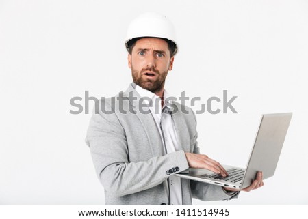 Confused bearded man builder wearing suit and hardhat standing isolated over white background, showing laptop computer