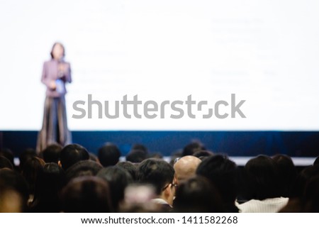 Blurry of woman speaker speech on stage in auditorium for shareholders' meeting or seminar event with projector and white screen, many business people listening on the conference. presenting concept