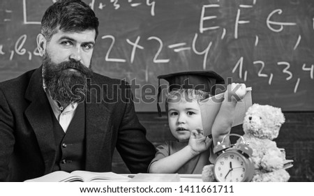 Father with beard, teacher teaches son, kid boy. Teaching kid concept. Teacher and pupil in mortarboard, chalkboard on background. Kid studies with teacher, near copybook, clock, toy.