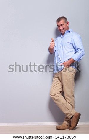 full length picture of a casual senior man standing with a hand in his pocket and showing thumbs up while smiling for the camera. on gray background