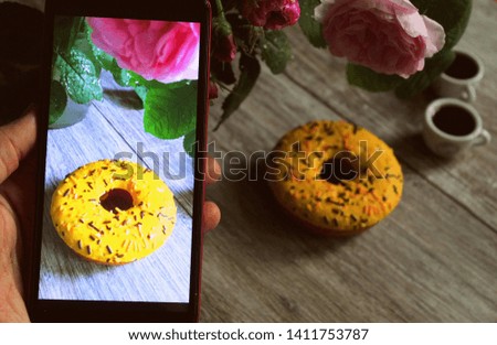 Phone in hand with a picture of a donut and coffee with flowers, breakfast on the table, selective focus.