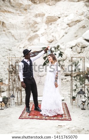 bride and groom in canyon outdoors. wedding ceremony. boho wedding arch. African man and Caucasian woman in boho wear are dancing on red carpet on the background of arch and decor