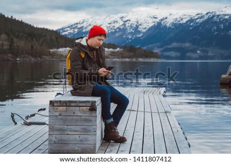 Traveler man looking into the phone. Tourist in yellow backpack sitting on a background of a mountain and a lake a wooden pier. Traveler walks, takes photo in mountains.