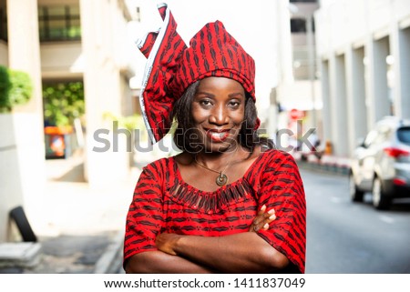 mature african woman standing outdoors head tied with a piece of loincloth watching the camera smiling.