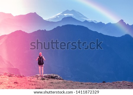 Girl traveler with backpack stands on a stone back to the viewer. She looks at the snow-capped top. Picturesque cloudless landscape with a rainbow. Wallpaper for concept of business thinking. climbing