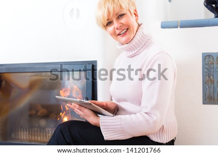 Quality of life - Older Woman or female Pensioner sitting at home in front of the furnace, writing emails on the tablet computer or reading a e-book