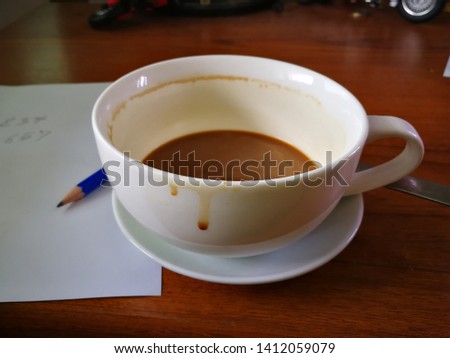 The white coffee mug has coffee stains on the desk. Blue pencil placed on a white sheet of paper. Brown wood table