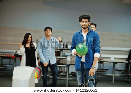 South asian man in jeans shirt standing at bowling alley with ball on hands. Guy is preparing for a throw. Friends support him.