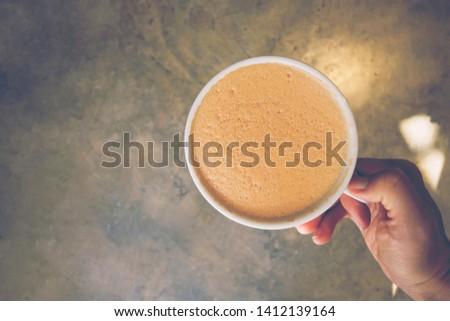 Hand holding cup of cappuсcino, gray concrete background with sunlight, toned. Top view, copy space