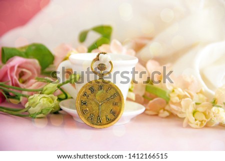 A coffee white cup next to it is a gold-colored decorative pocket watch, fresh pink flowers and a white cloth. Concept of congratulatory or background about time, emblazoned card. Copy spacy