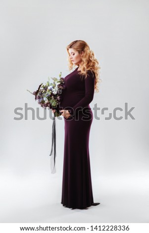 Portrait of young pregnant woman with big flower in her hand. Beautiful model posing in dark dress and holding her stomach at white studio background. Beautiful and happy pregnancy concept