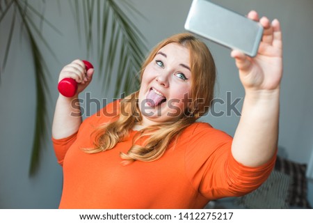 Chubby woman sport at home standing holding dumbbell taking selfie photos showing tongue playful