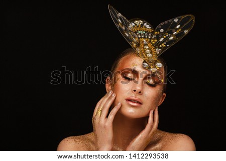 Portrait of a beautiful woman with expressive creative make-up in bronze and with a decoration on her head in the form of a fly. Studio photo session. Black background