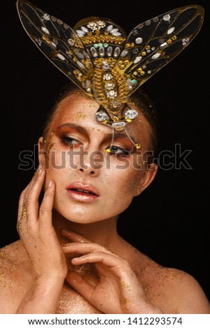Portrait of a beautiful woman with expressive creative make-up in bronze and with a decoration on her head in the form of a fly. Studio photo session. Black background