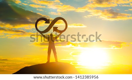 A silhouette of a man holds an infinity symbol on top of a mountain with a sunset background. Concept idea, life, success, business.