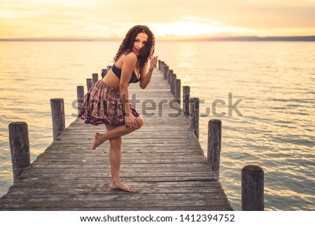 
Happy woman enjoying the sunset at the lake on her vacation. Woman in summer outfit stands laughing on the boardwalk of Lake Neusiedl with curly dark hair. Footbridge running in the picture