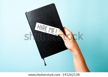 Hand holding a black notebook/book with a label "Hire me", on blue background. Hiring concept.