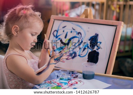 portrait of a beautiful little girl who diligently paints her hands with colored paints, in a kindergarten drawing class a child