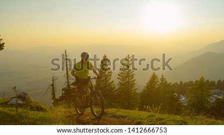 LOW ANGLE, LENS FLARE: Active male tourist pedalling a bright green electric bike uphill at sunrise. Happy young Caucasian man riding his mountain bike up gravel path on a beautiful summer evening.