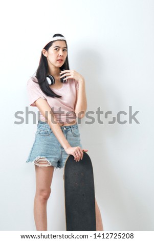 Beautiful asian woman model in white t-shirt and denim shorts  holding skateboard and smiling  on white wall background