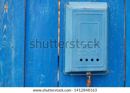 An old blue Soviet mailbox with the inscription Mail and a rusty lock hanging on a rural wooden blue fence. Close-up.