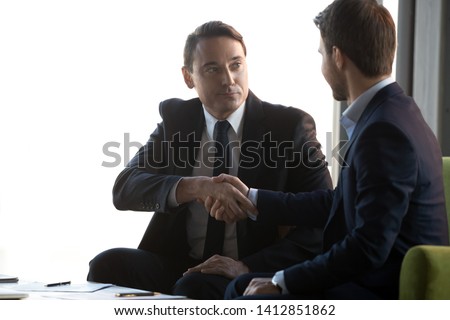 Serious businessmen shake hands sitting in office after successful negotiations, signing contract closing deal, business partners handshake greeting with partnership, making agreement at briefing