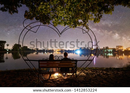 Back view of man and woman sitting on a bench near bonfire on the shore near lake under tree. Couple enjoying beautiful view of evening sky full of stars, Milky way and luminous town on background