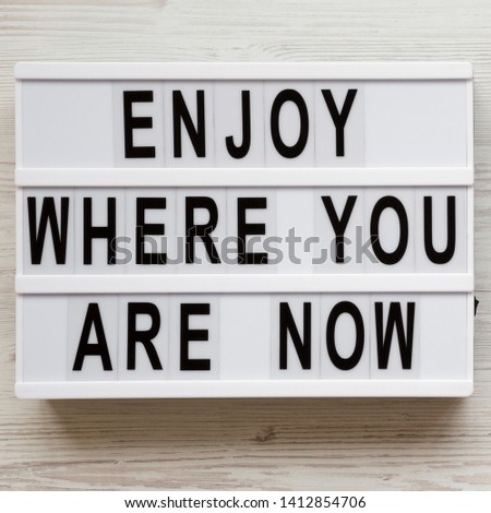 'Enjoy where you are now' words on a modern board over white wooden background, overhead view. Top view, flat lay, from above.