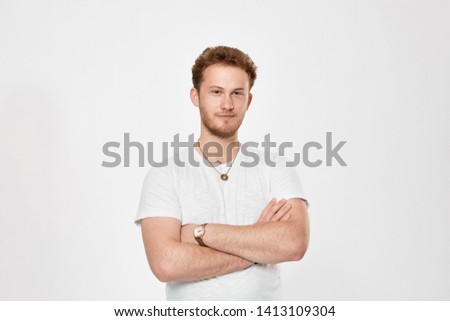 Portrait of a confident handsome young man with a beard infront of white bakground. Model in a white t-shirt smiles at the camera and keeps arms crossed.