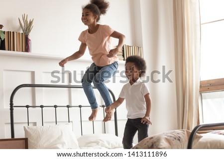 Happy cute active little african american kids boy and girl jumping on bed laughing together, two funny small energetic mixed race children brother with sister having fun play active game in bedroom