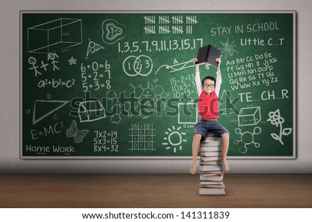 Asian boy lifting a book while sitting on stack of books in classroom