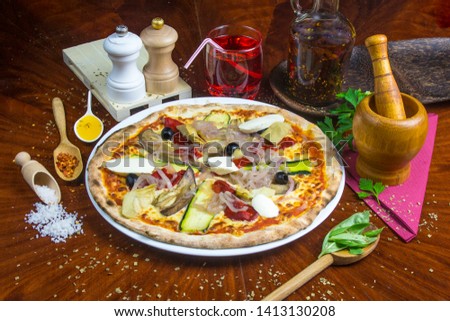 Bio pizza vegeterian with artichoke, aubergine, mozzarella, courgette, sweet pepper, olive, onion. Presented with spicy oil and several decorative elements. Served on a round wooden table.