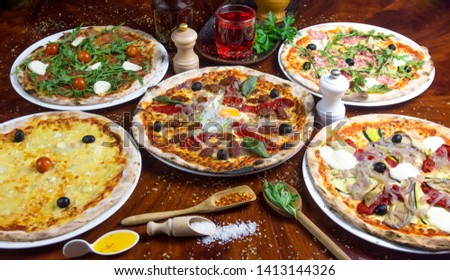 5 bio pizza: cheese, tomato, eastern, salad, vegetarian, mozzarella, romana. Presented with spicy oil and several decorative elements. Served on a round wooden table.