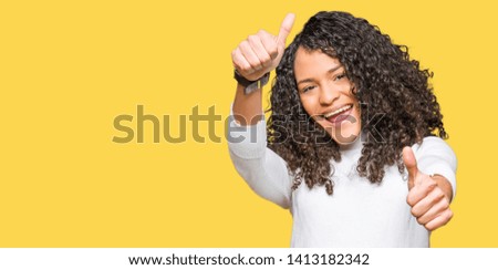 Young beautiful woman with curly hair wearing turtleneck sweater approving doing positive gesture with hand, thumbs up smiling and happy for success. Looking at the camera, winner gesture.