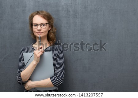 Studio waist-up portrait of calm caucasian fair-haired young woman with glasses, smiling slightly and looking at camera, holding folder and pen in hands, against gray background, copy space on right