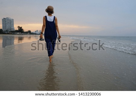Summer vacation time theme woman wearing hat and blue overalls holding flip flop sandal while walking barefoot by the seaside with blurred background of sand and sea at sunset moment. (space for text)