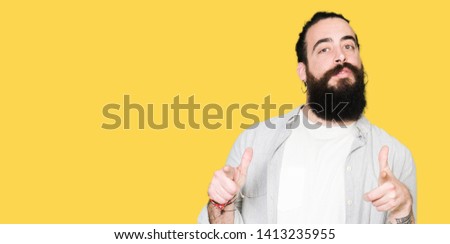 Young man with long hair, beard and earrings pointing fingers to camera with happy and funny face. Good energy and vibes.