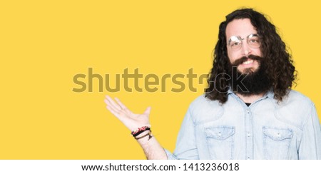 Young hipster man with long hair and beard wearing glasses smiling cheerful presenting and pointing with palm of hand looking at the camera.