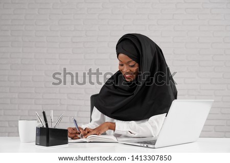 Front view of beautiful female wearing black hijab smiling and writing while sitting at table at office. Young african woman smiling and enjoying working process in company. Concept of business.