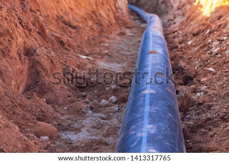 A black plastic pipe with a blue strip to feed water lies in a dug trench. Royalty-Free Stock Photo #1413317765