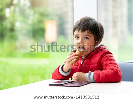 Happy child licking an ice cream cone, Smiling kid sitting alone watching cartoon on mobile phone and eating an ice cream, Relaxing preschool boy sitting at the table in a cafe with blurry background