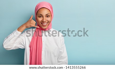 Happy young Muslim female model gestures with fingers, says call me, smiles broadly, wears pink hijab and white shirt, models over blue background, copy space for advertising content. Body language