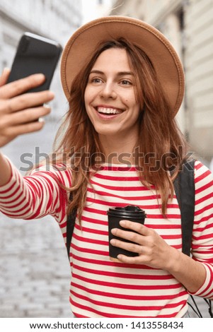 Pretty glad traveler clicks selfie photo, makes new pictures, uses cell phone and application, enjoys leisure outdoors, drinks aromatic coffee from disposable cup, wears headgear and striped jumper.