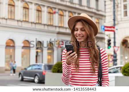 Smiling young European woman enjoys recreation time, watches interesting video on website, wears stylish headgear and striped sweater, has joyful expression, poses over urban setting, chats online