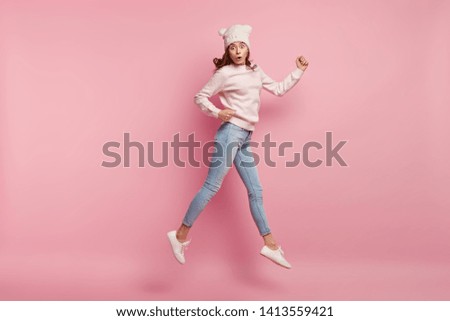 Impressed young European woman jumps in air, makes movements over pink background, wears hat, jumper and jeans, being speechless, gasps from shock. People, human emotions and feelings concept