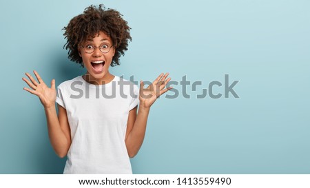 Curly optimistic woman raises palms from joy, happy to receive awesome present from someone, shouts loudly, dressed in casual white t shirt, isolated on blue background. Excited Afro female yells Royalty-Free Stock Photo #1413559490