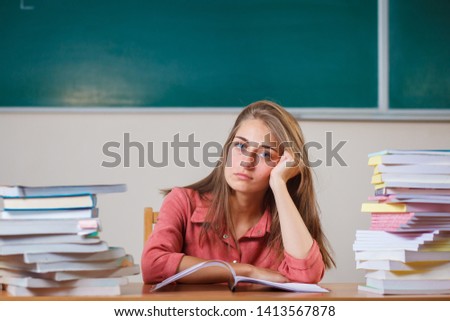 Teacher surrounded by books sitting in school classroom.Emotional teacher at the table. Copy space. Funny photo. Beautiful young girl.Many study tasks