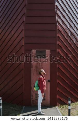 Urban lifestyle concept. Full length side on portrait of young smiling hipster guy staying on red building door background and enjoying sun shine