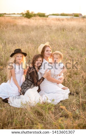 A young mother with her daughters and an aunt with blond hair in white dresses at sunset in the summer in a field of countryside. Several girls from the same family in the summer in the field

