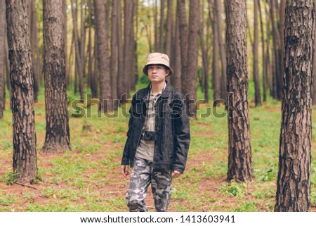 Asia man wears shirt and denim jacket, hat, and camouflage pants are walking and taking photos at the forest.